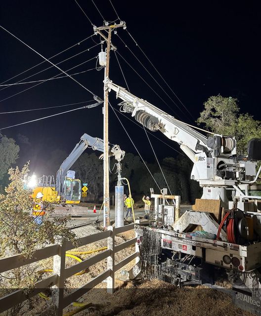 Digger derrick and air-hammer rock drilling at night beside a power pole