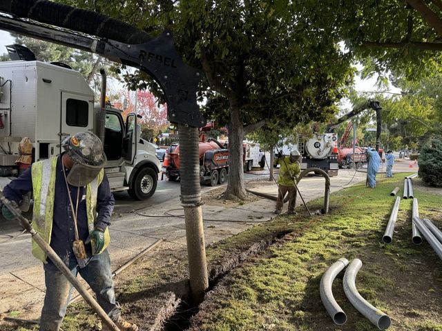 Crew member vacuum-excavating a trench with conduit sweeps staged nearby